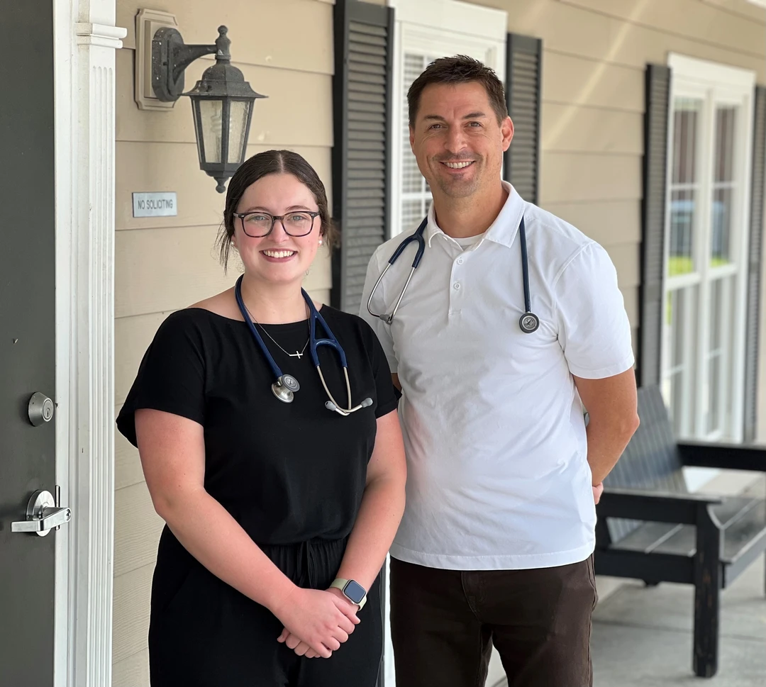Two healthcare professionals, one in a black outfit with a stethoscope and the other in a white polo, stand outside a building.